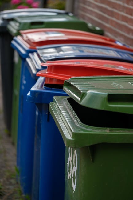 A row of large, rectangular plastic rubbish bins positioned along a pavement beside a brick wall. The bins are in various colors, including green, blue, red, and dark green, each with a hinged lid on