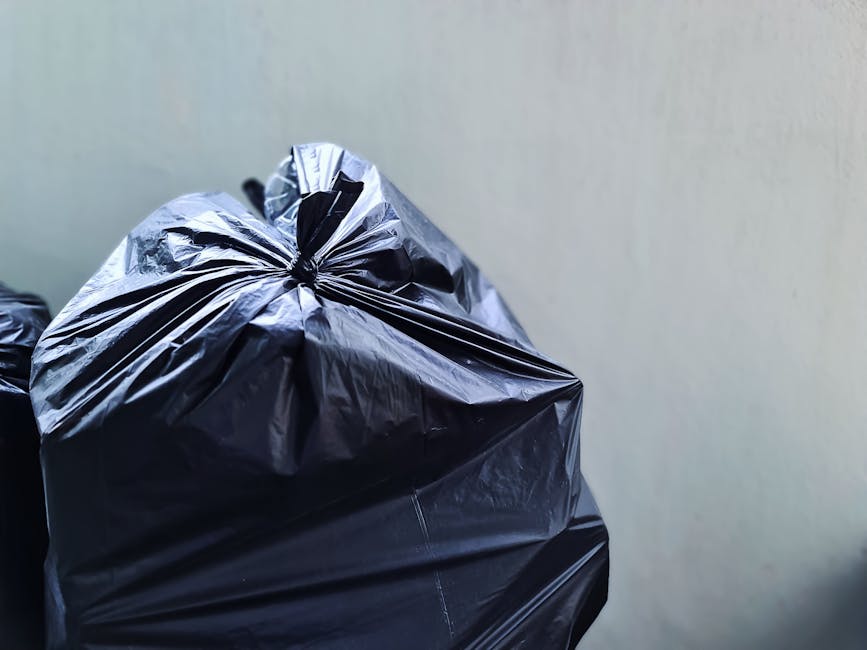 Close-up view of several black plastic rubbish bags filled with waste, tied at the top with the plastic material appearing shiny and crinkled. The bags are stacked against a plain, light-colored wall,