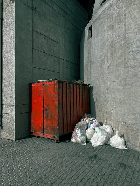 Close-up view of several black plastic rubbish bags filled with waste, tied at the top with the plastic material appearing shiny and crinkled. The bags are stacked against a plain, light-colored wall,